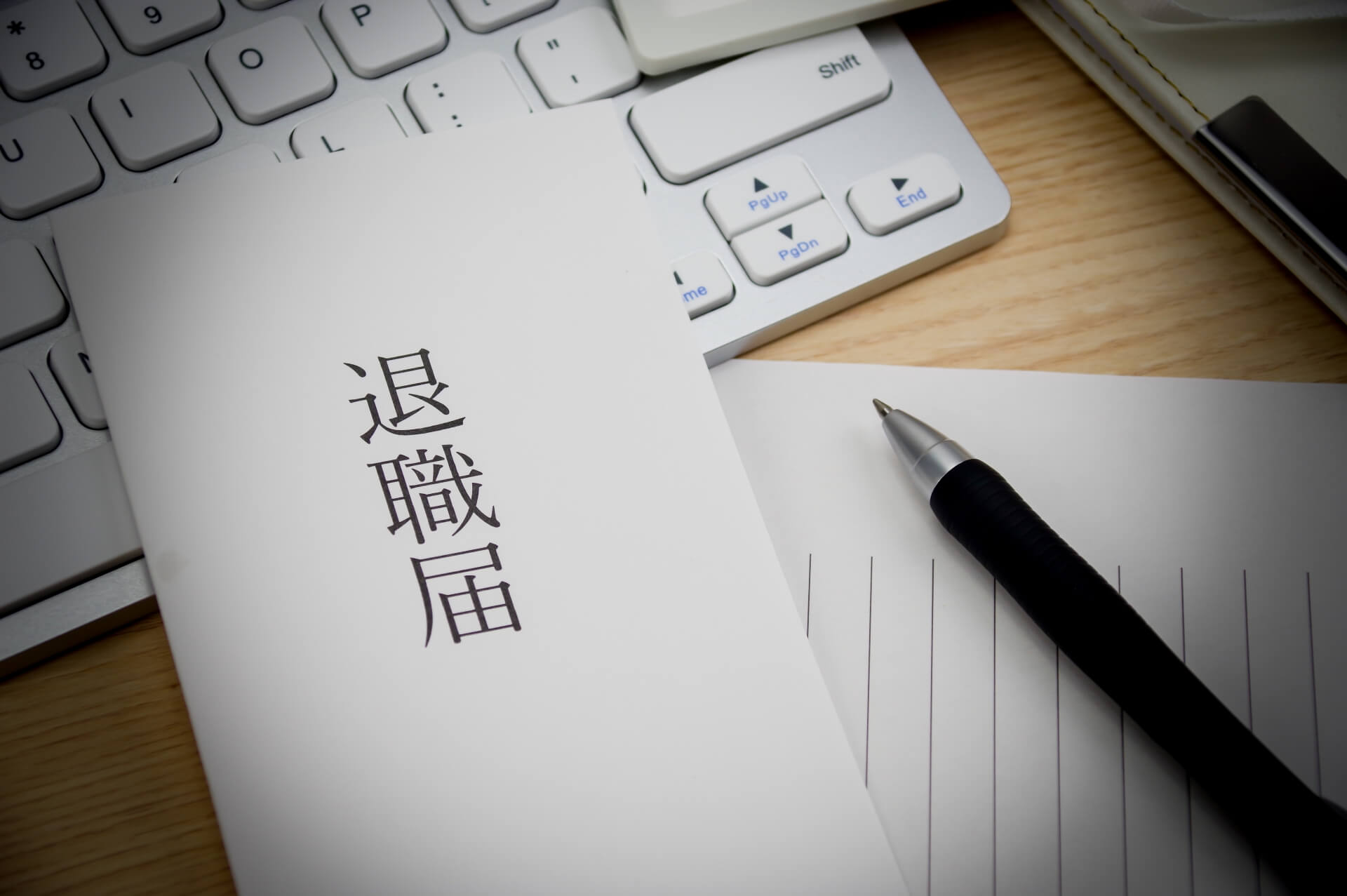 退職届とペン、パソコンのキーボードが並ぶ写真。美容師がお店や会社を辞めたいのに辞めさせてもらえないときのイメージ。