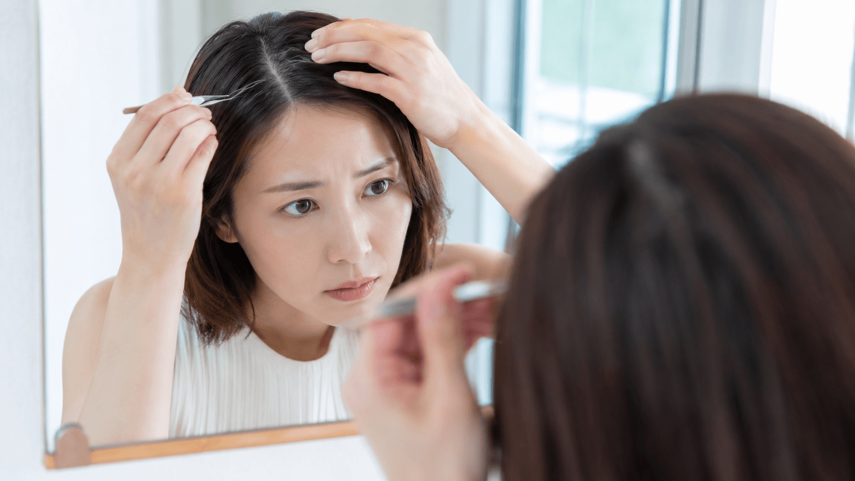 分け目に白髪を見つけて毛抜きに手を伸ばそうとする30代女性のシーン